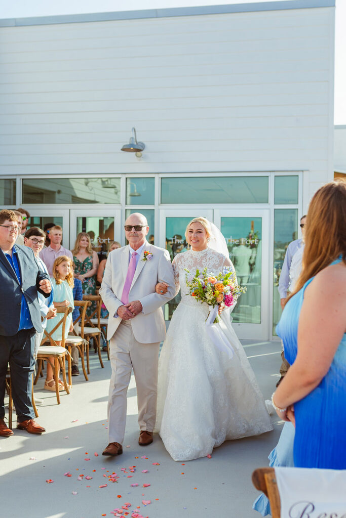 The bride walks down the aisle with a family member, carrying a vibrant bouquet as guests look on.