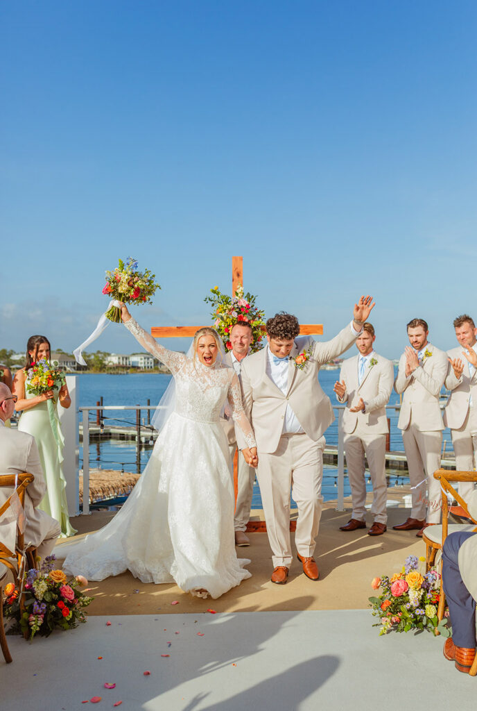 The newly married couple celebrates at the altar, raising joined hands in excitement as their wedding party applauds.