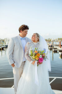 The newlyweds share a portrait by the marina, smiling at one another as the bride holds a colorful bouquet.