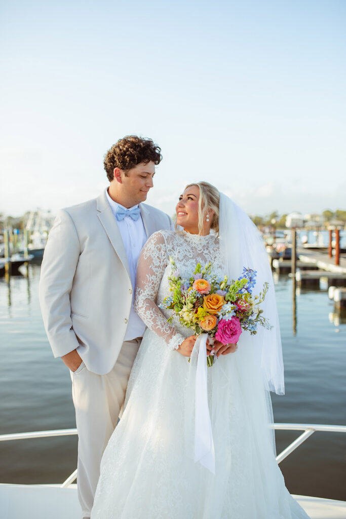 The newlyweds share a portrait by the marina, smiling at one another as the bride holds a colorful bouquet.