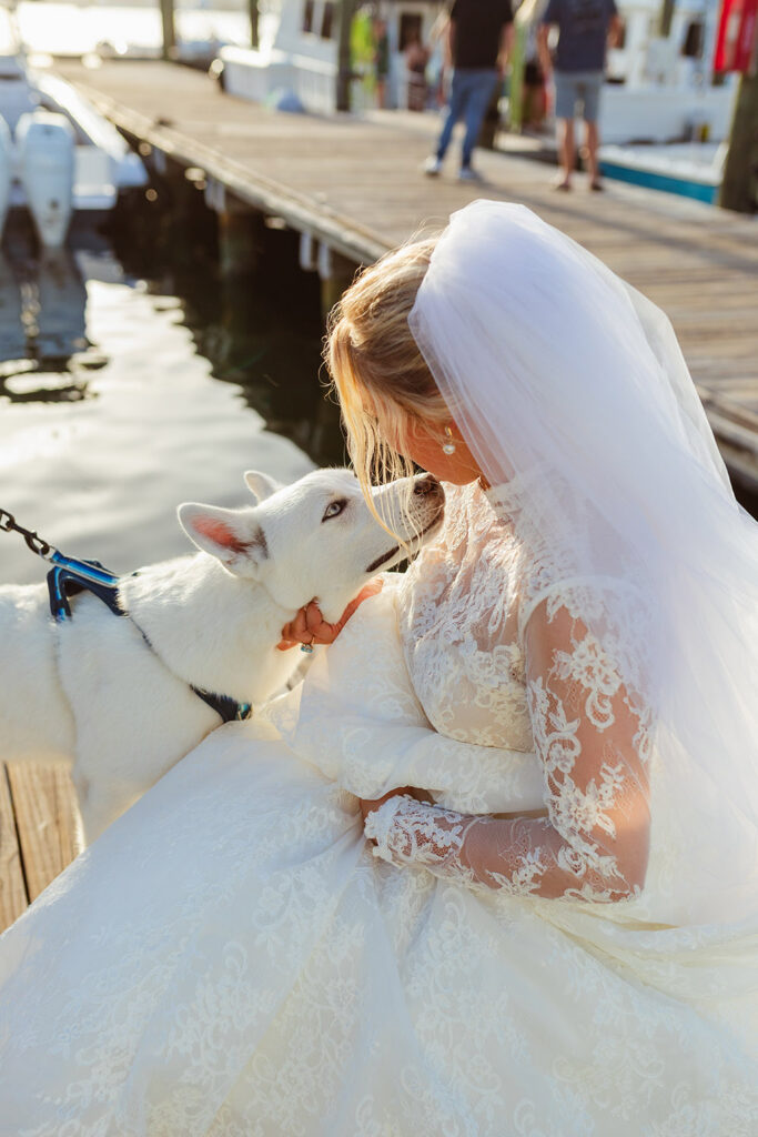The bride kneels on a dock to greet a white dog, sharing a sweet moment before the reception.