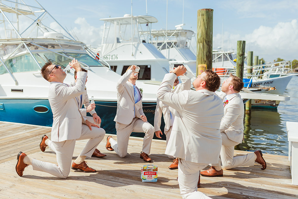 Groomsmen kneel on a marina dock, taking celebratory shots beside docked boats before the ceremony.