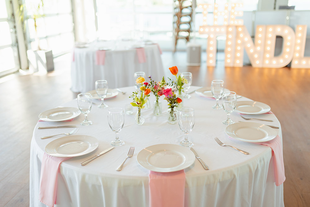 A reception table is set with white place settings, blush napkins, and bud vase floral centerpieces in the ballroom.