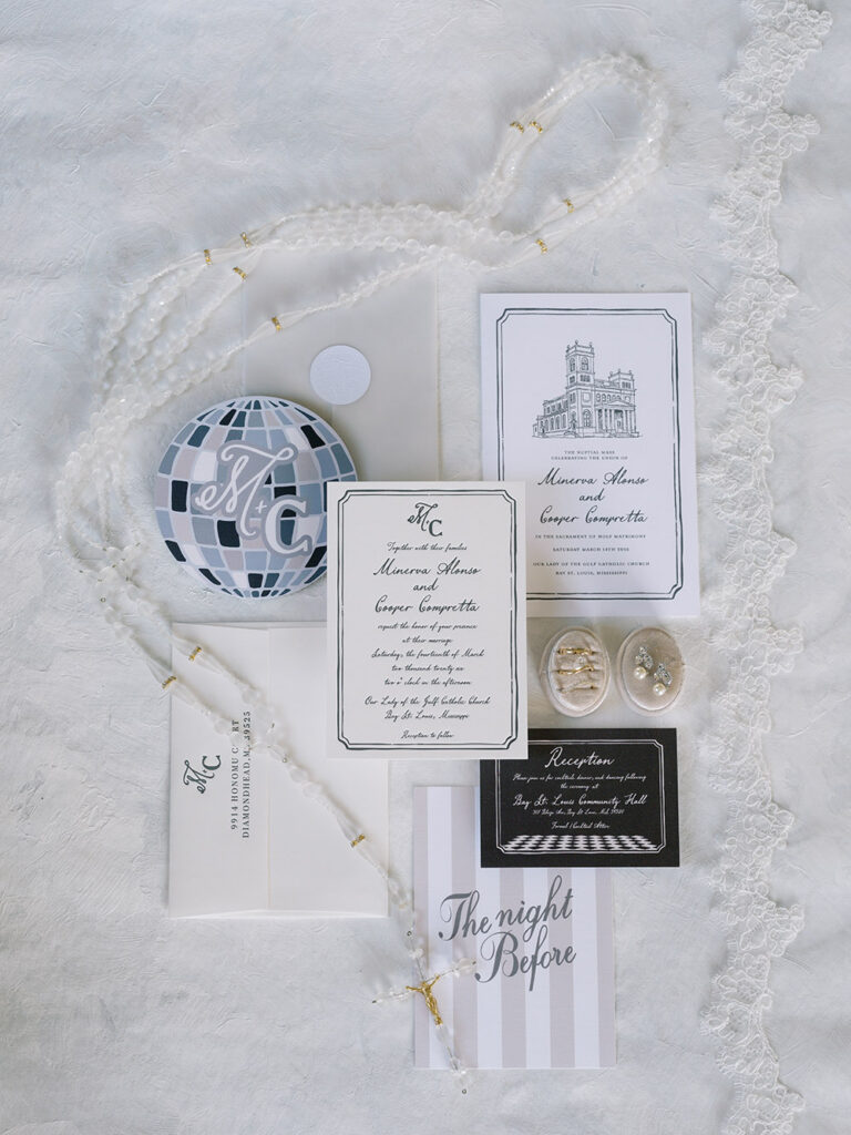 Flat lay of wedding details featuring custom invitations, vow booklet, rings, rosary, lace veil, and a disco ball-inspired monogram design.