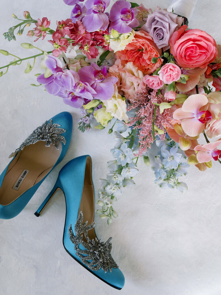 Flat lay of vibrant bridal bouquet with orchids, roses, and ranunculus beside blue Manolo Blahnik heels adorned with crystal embellishments.