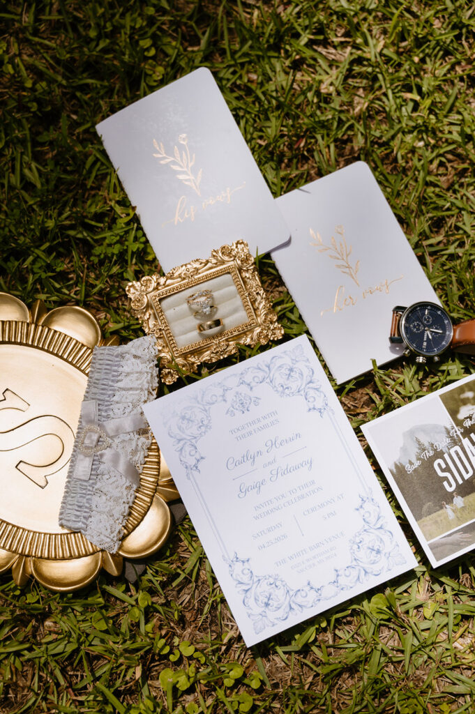 Flat lay of wedding details including invitation suite, rings, heirloom vow books, watch, lace garter, and save-the-date card arranged on grass.