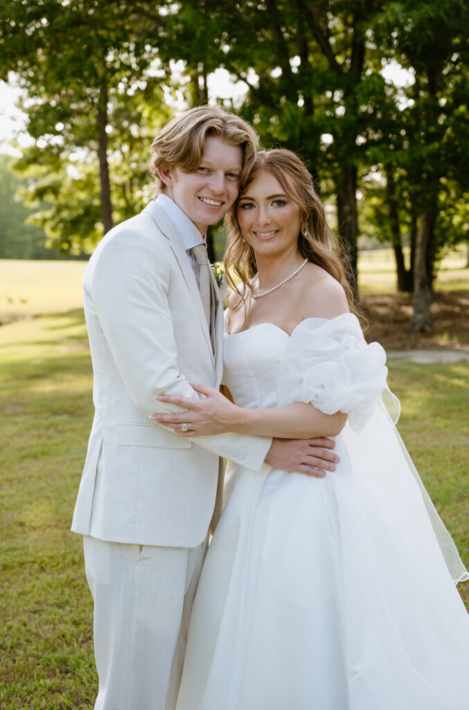 Bride and groom embrace outdoors, smiling at the camera after their ceremony.