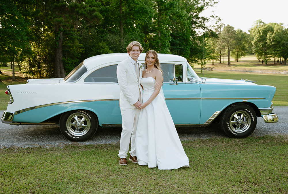 Bride and groom pose in front of a vintage blue-and-white classic car.