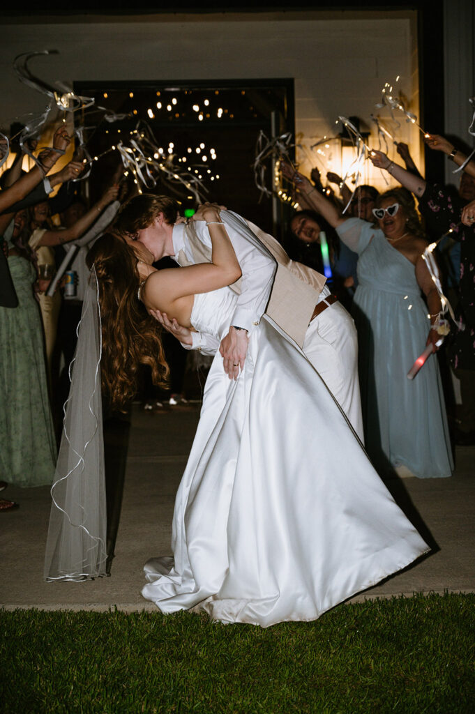 Bride and groom share a dipped kiss during a sparkler exit surrounded by cheering guests.