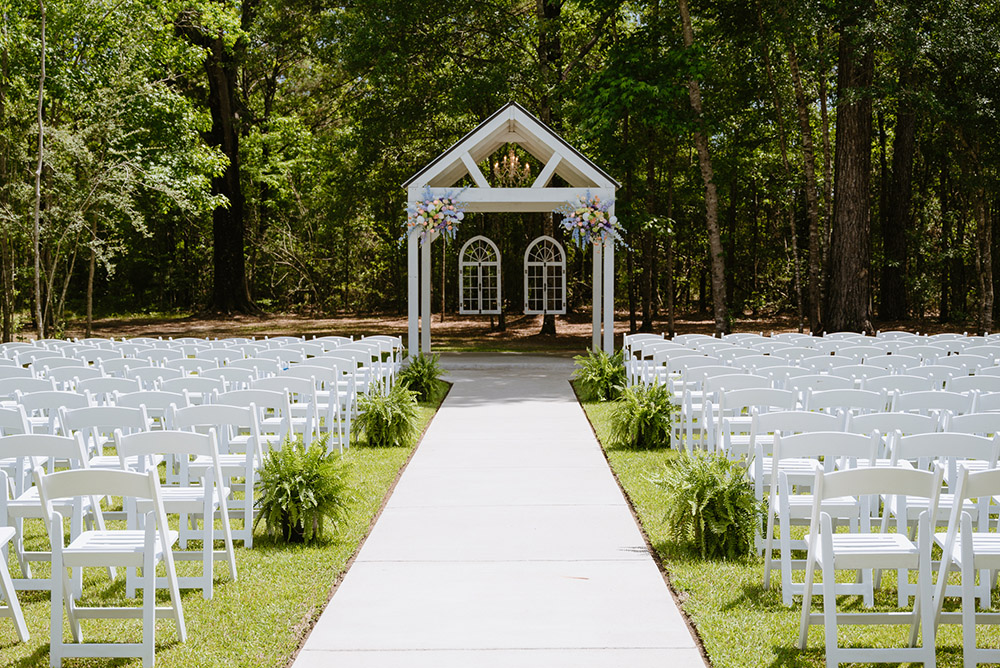 Outdoor wedding ceremony space with white chairs lining a long aisle leading to a flower-adorned white arbor.