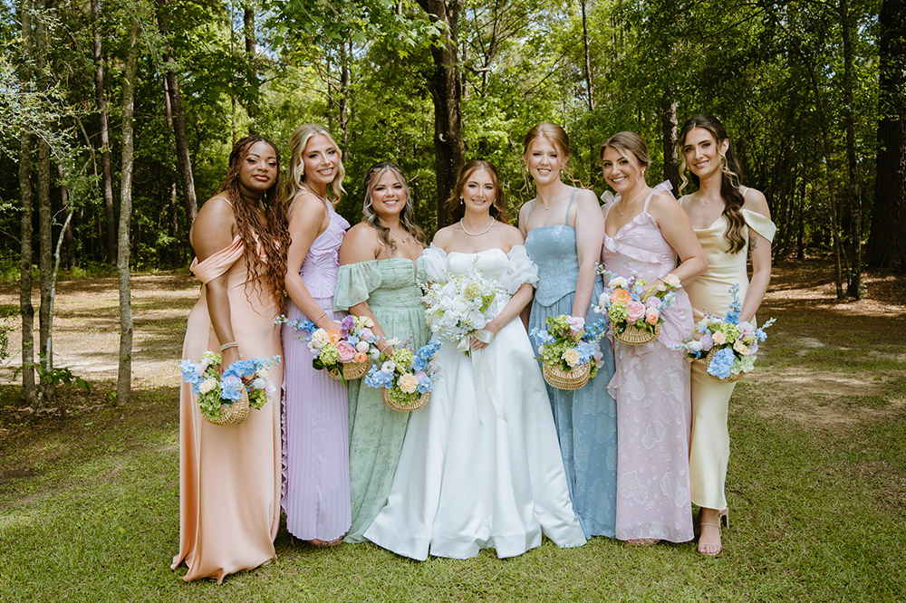 Bride poses with six bridesmaids wearing pastel dresses and carrying woven basket floral arrangements.