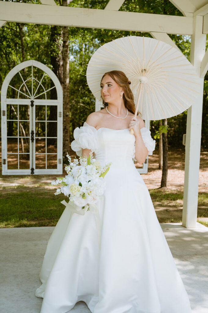 Bride in an off-the-shoulder satin gown holds a white bouquet beneath a parasol at the ceremony arbor.