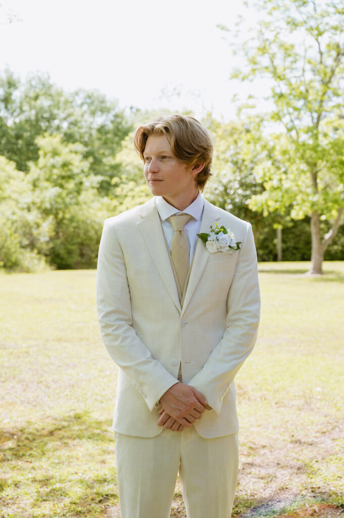 Groom in a light cream suit stands outdoors before the ceremony with hands clasped.