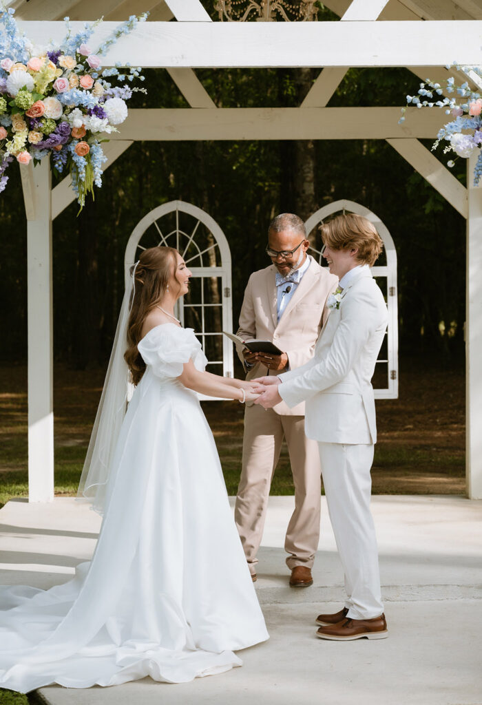 Bride and groom hold hands as they exchange vows beneath a flower-adorned outdoor arbor.