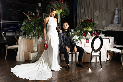 A woman in a white gown stands beside a seated man in a black suit at an elegant table decorated with flowers and greenery, set in a formal event space.