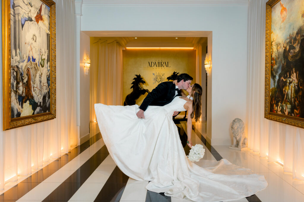 Groom in black tuxedo kisses bride in white gown on tiled hallway with art, statues, and “ADMIRAL” sign before Gulf South wedding.