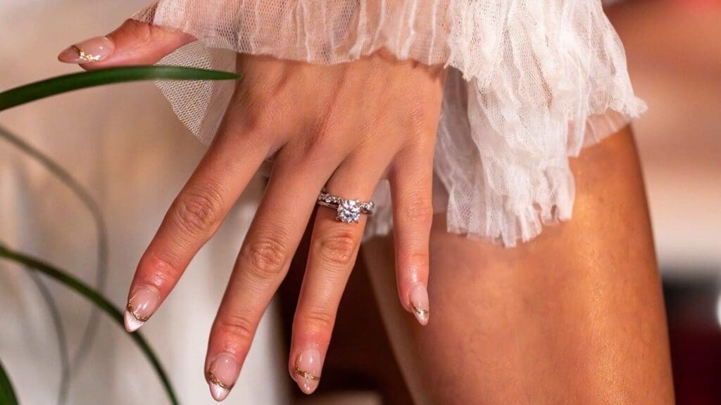 Manicured hand with diamond engagement ring, in white frilly sheer fabric, near a green plant.