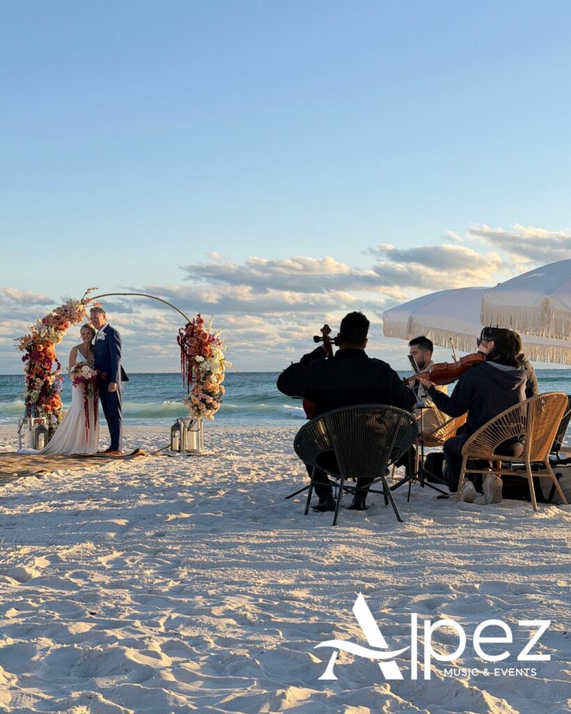 Couple under floral arch at Gulf South beach wedding, string quartet nearby, ocean view, bright scene, Alpez logo in corner.