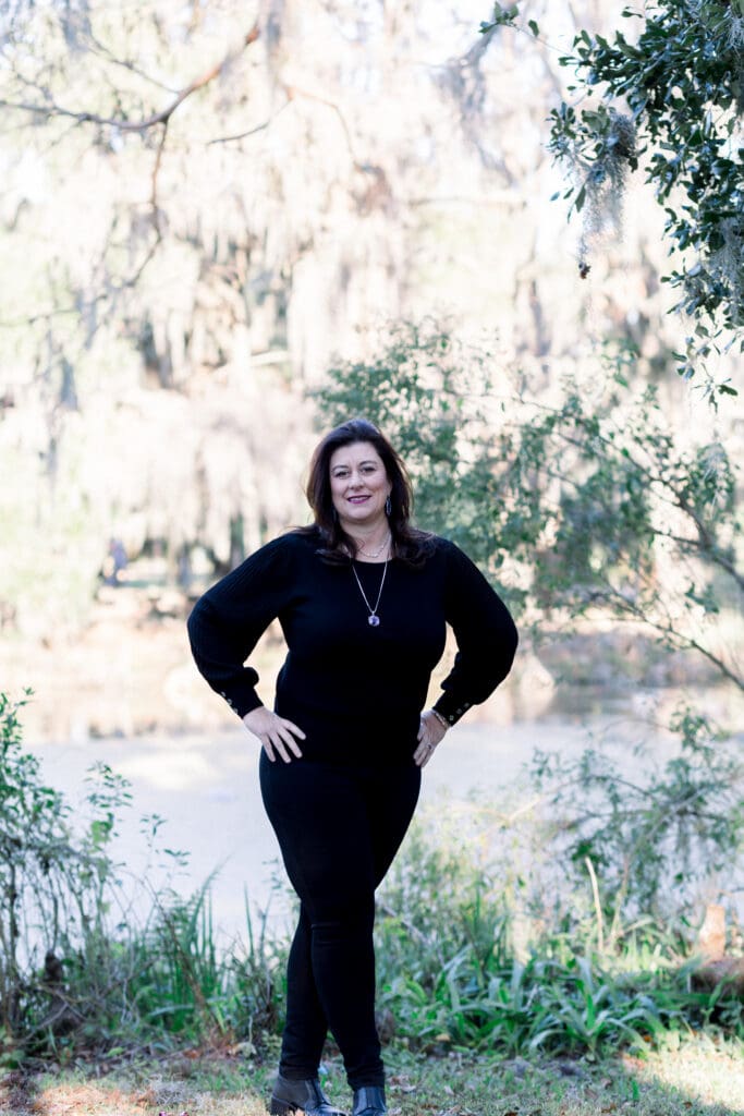Woman in a black outfit stands by trees and a river, smiling with hands on hips—effortless Louisiana wedding inspiration.