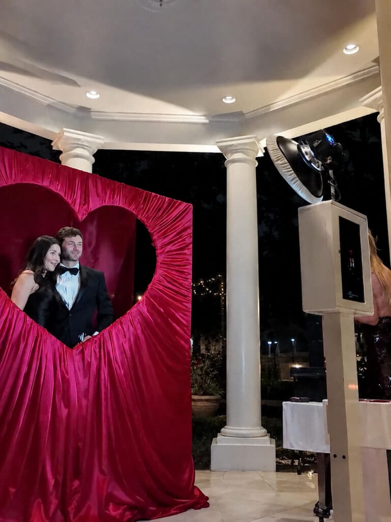 Couple posing in a red heart-shaped frame at an outdoor New Orleans wedding, with photo booth and white columns behind them.