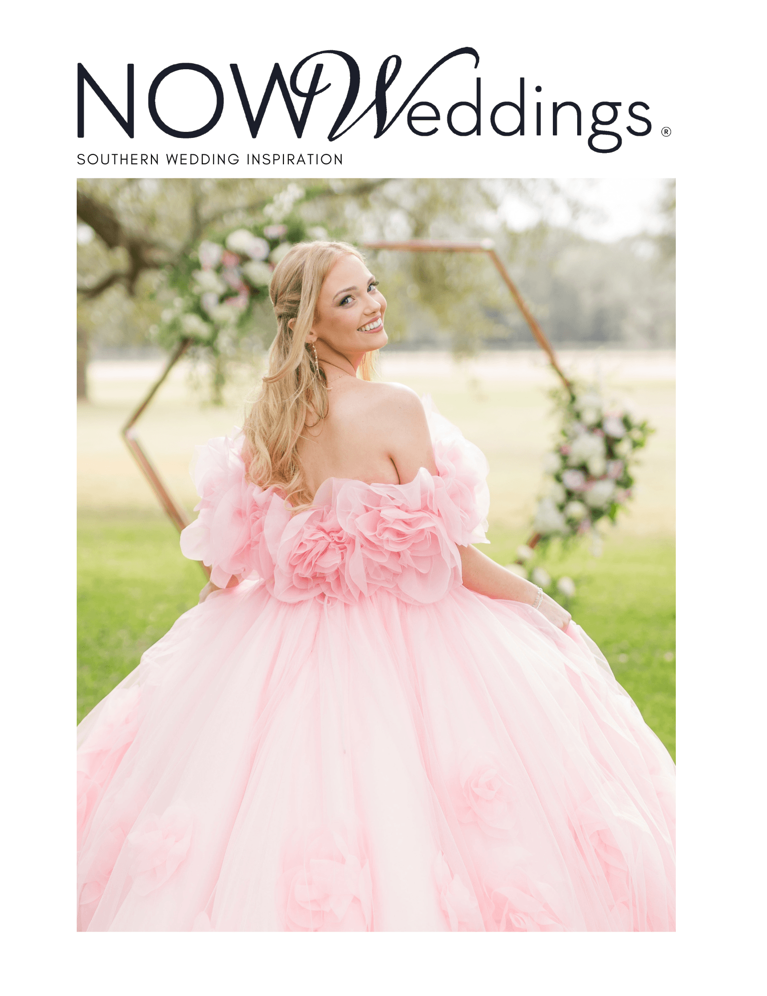 Bride in a pink floral gown smiles outdoors before a wooden hexagonal arch adorned with flowers.