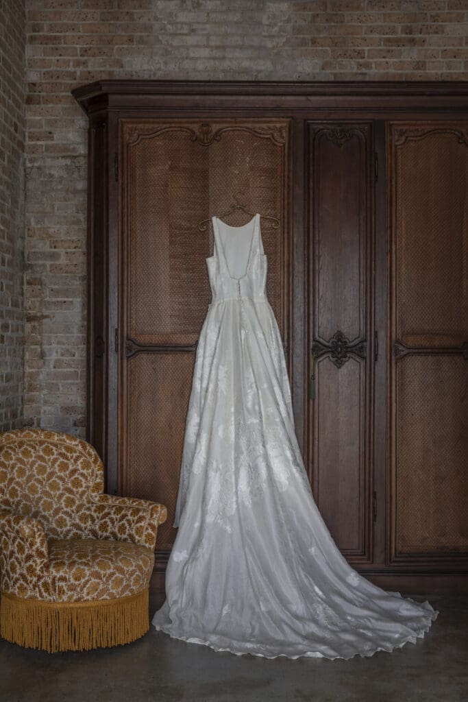 White floral wedding dress on wooden armoire next to gold fringe chair, brick wall backdrop—classic Louisiana wedding charm.