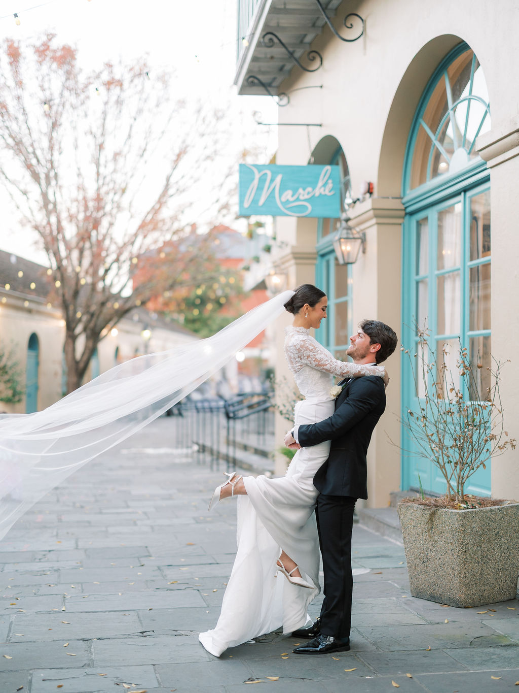 Groom lifts bride outside blue-doored Marche in Louisiana; her veil flows as they smile, trees with autumn leaves behind them.