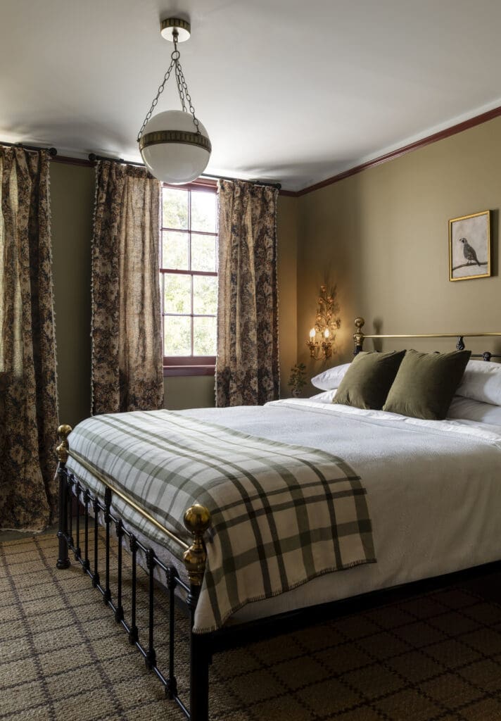 Cozy bedroom with metal bed, white bedding, plaid blanket, floral curtains, and vintage light fixture on olive green wall.