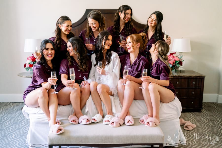 Eight women in matching purple pajamas laugh on a bed with champagne, flowers, and a wooden headboard in the background.