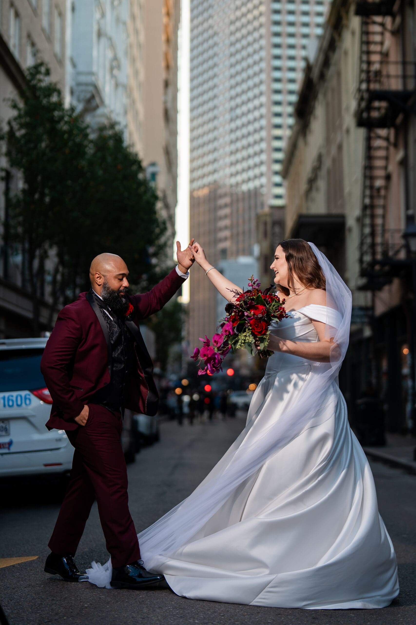 Bride in white gown and groom in burgundy suit high-five on a city street, celebrating a lively Louisiana wedding.