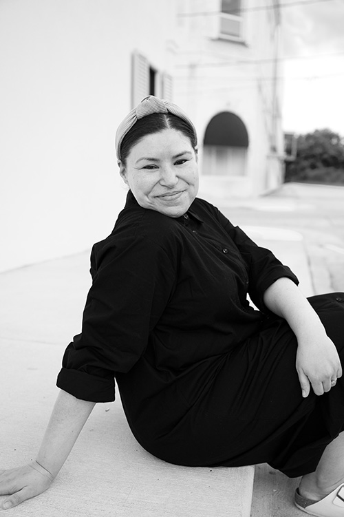 A smiling woman in a black outfit sits outdoors for a casual black-and-white portrait, wearing a knotted headband and glasses.