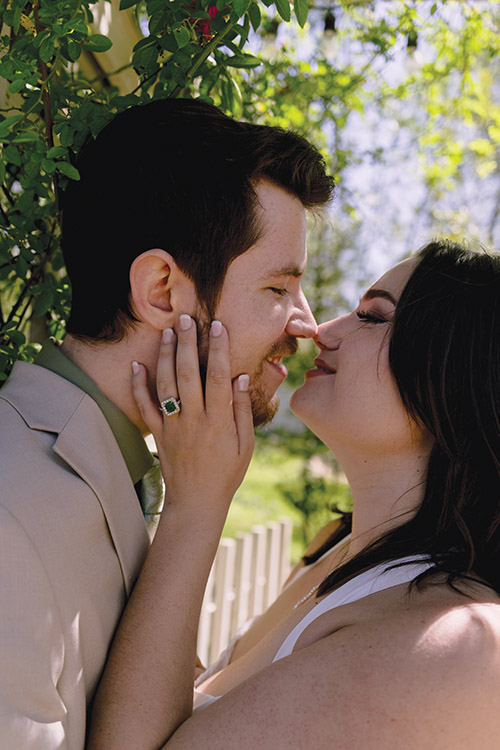 A close-up engagement portrait captures the couple smiling nose-to-nose beneath blooming wisteria as the bride-to-be shows off her emerald-cut engagement ring.