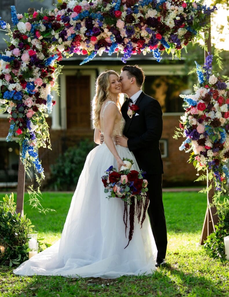 Bride and groom under a vibrant floral arch outdoors; he kisses her cheek as she smiles, holding a bright bouquet.