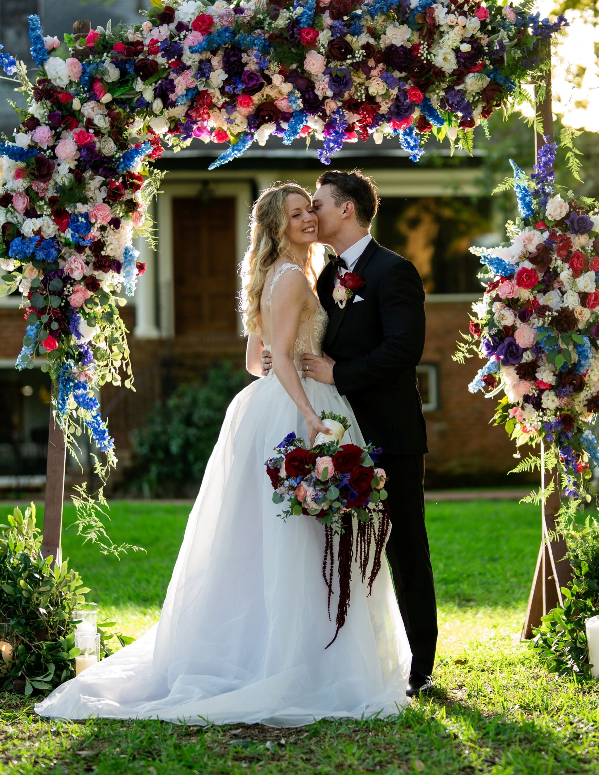 Bride and groom under a vibrant floral arch outdoors; he kisses her cheek as she smiles, holding a bright bouquet.