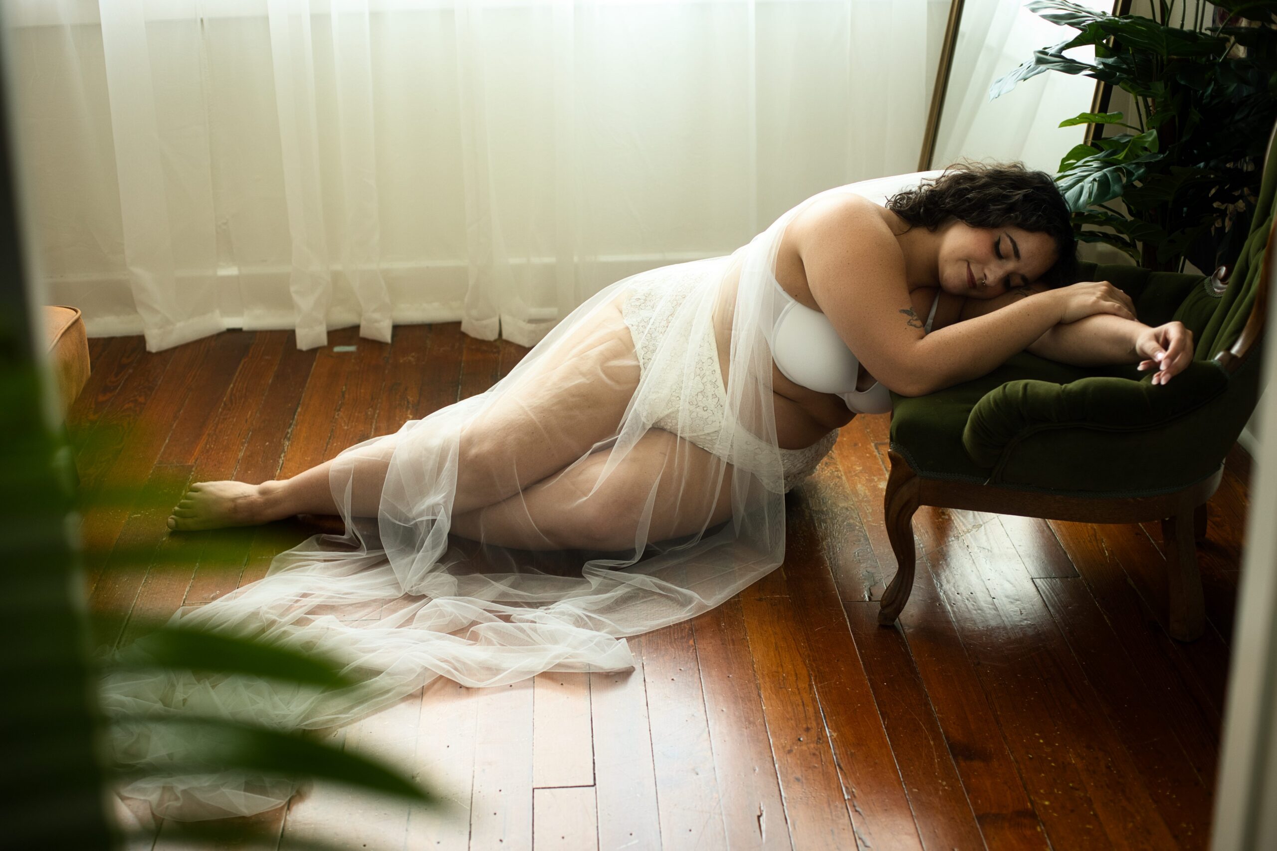 Woman in white lingerie and sheer robe leans on green chair by windows with sheer curtains, surrounded by plants.
