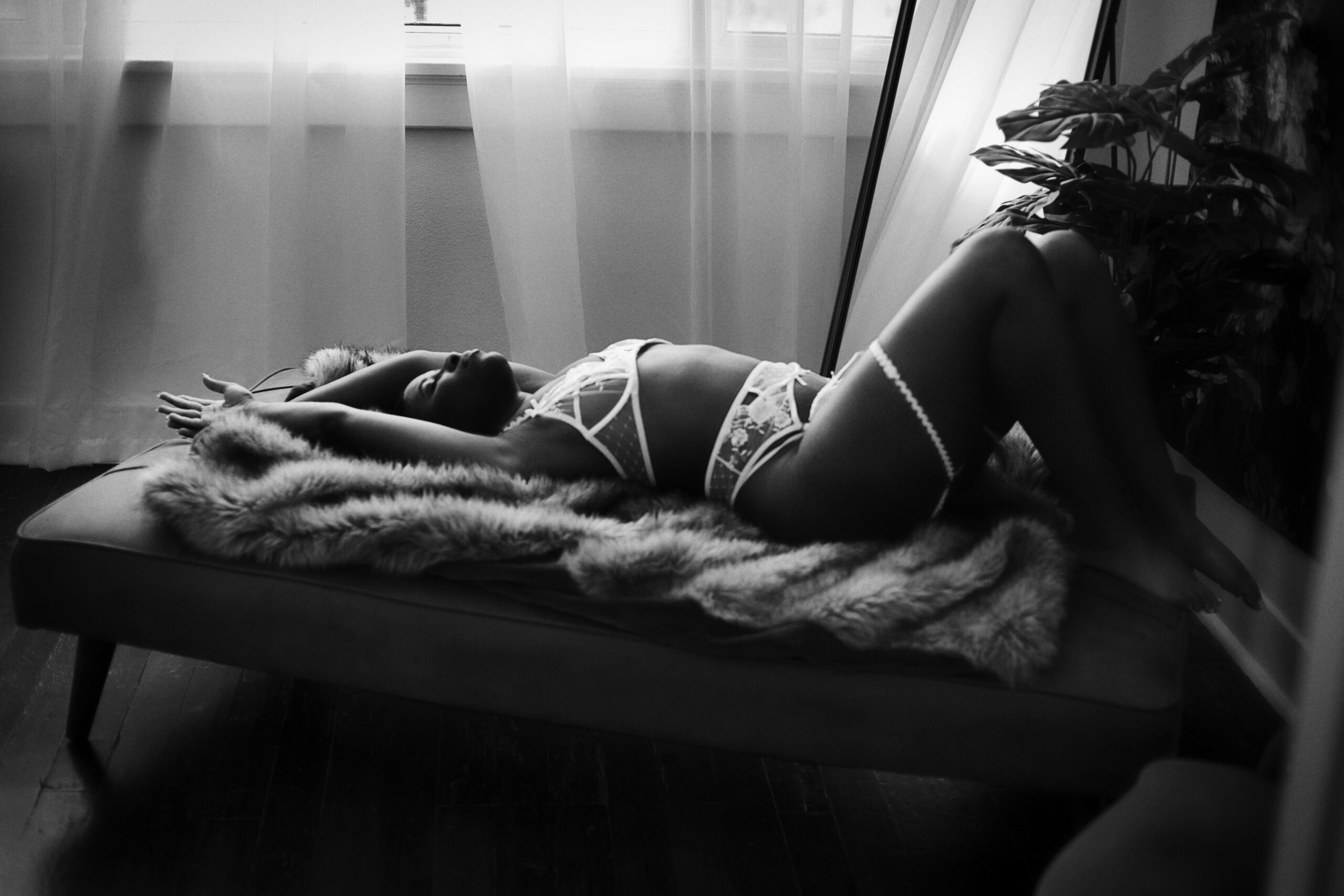 Woman in white lingerie reclines on a fur-covered chaise lounge in a softly lit room with sheer curtains and a plant.