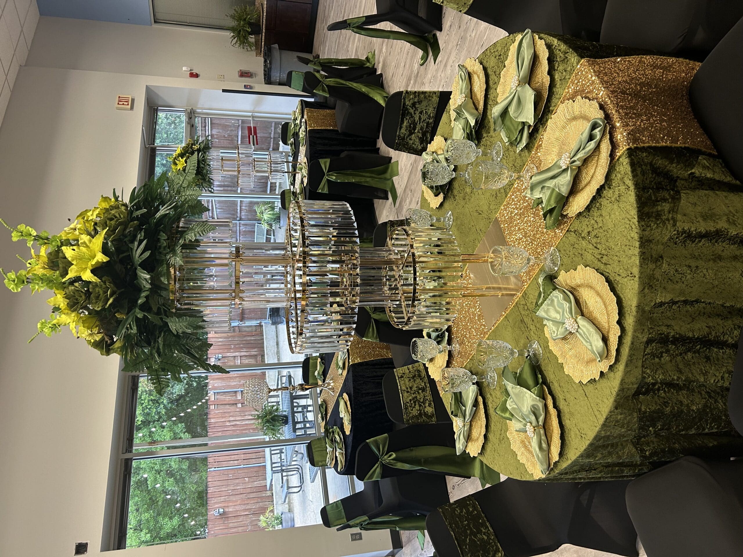 Elegant round table for six with gold plates, green napkins, foliage centerpiece, and black chairs with green sashes by large windows.