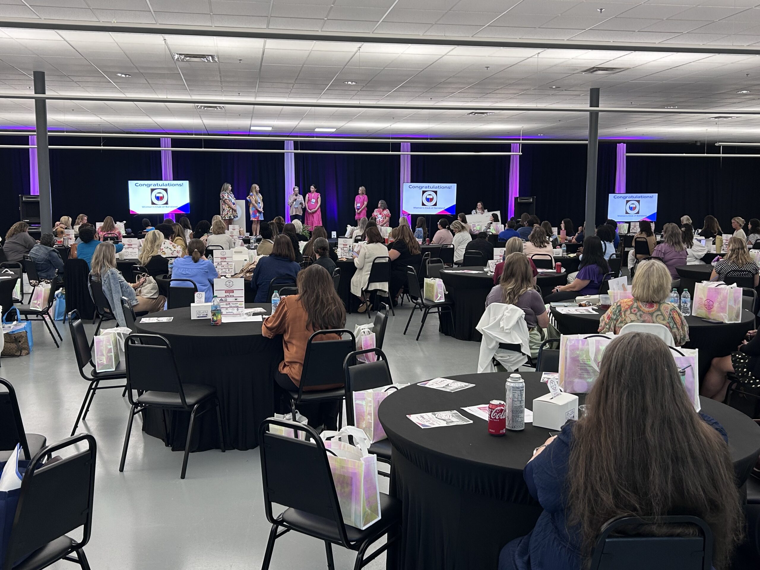 Attendees at round tables face a stage with five speakers; colorful logo on screens, purple lighting fills the large room.