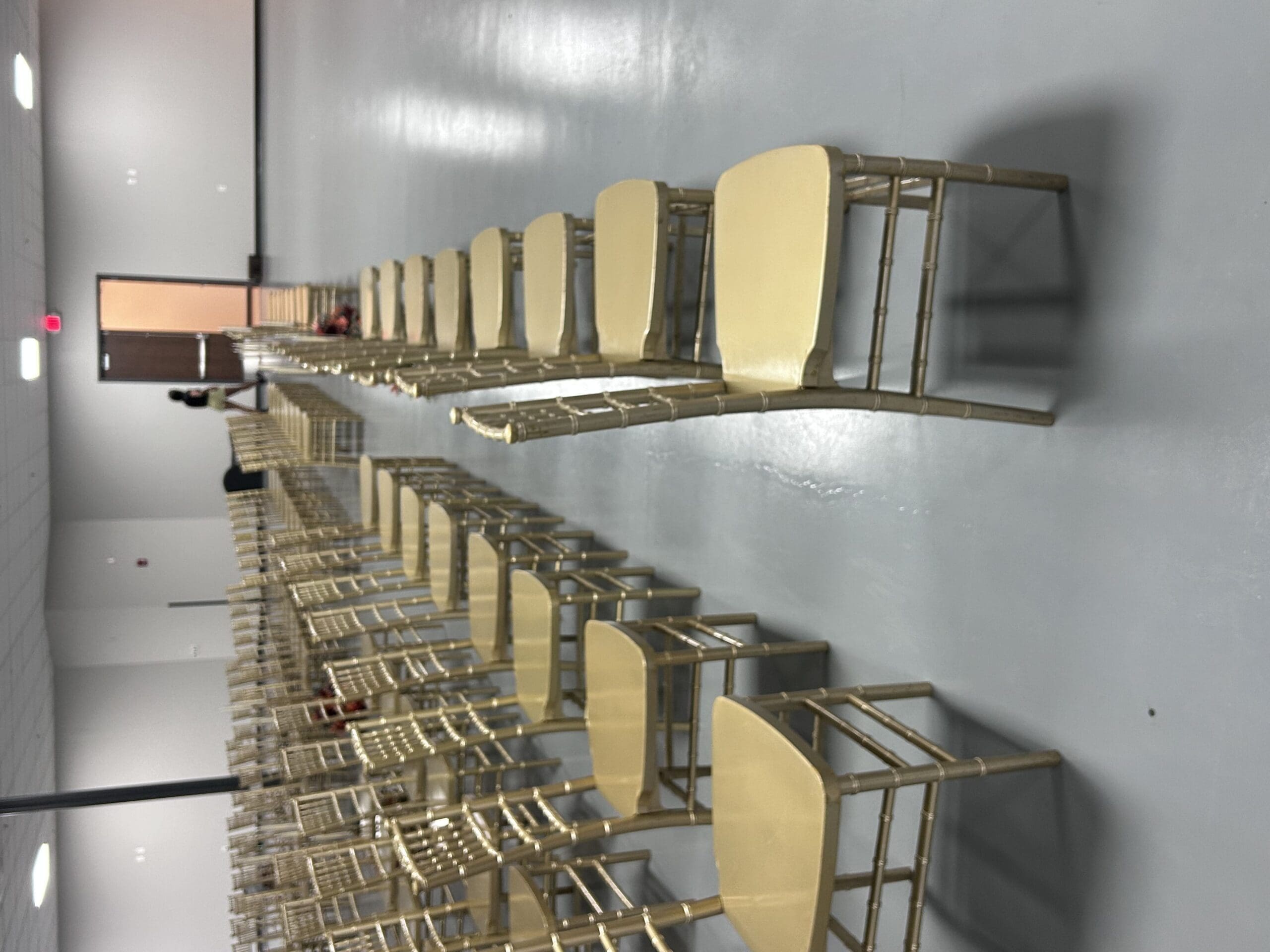 Gold chairs in rows fill a large, empty room with a shiny gray floor; one person stands near a door, ready for an event.