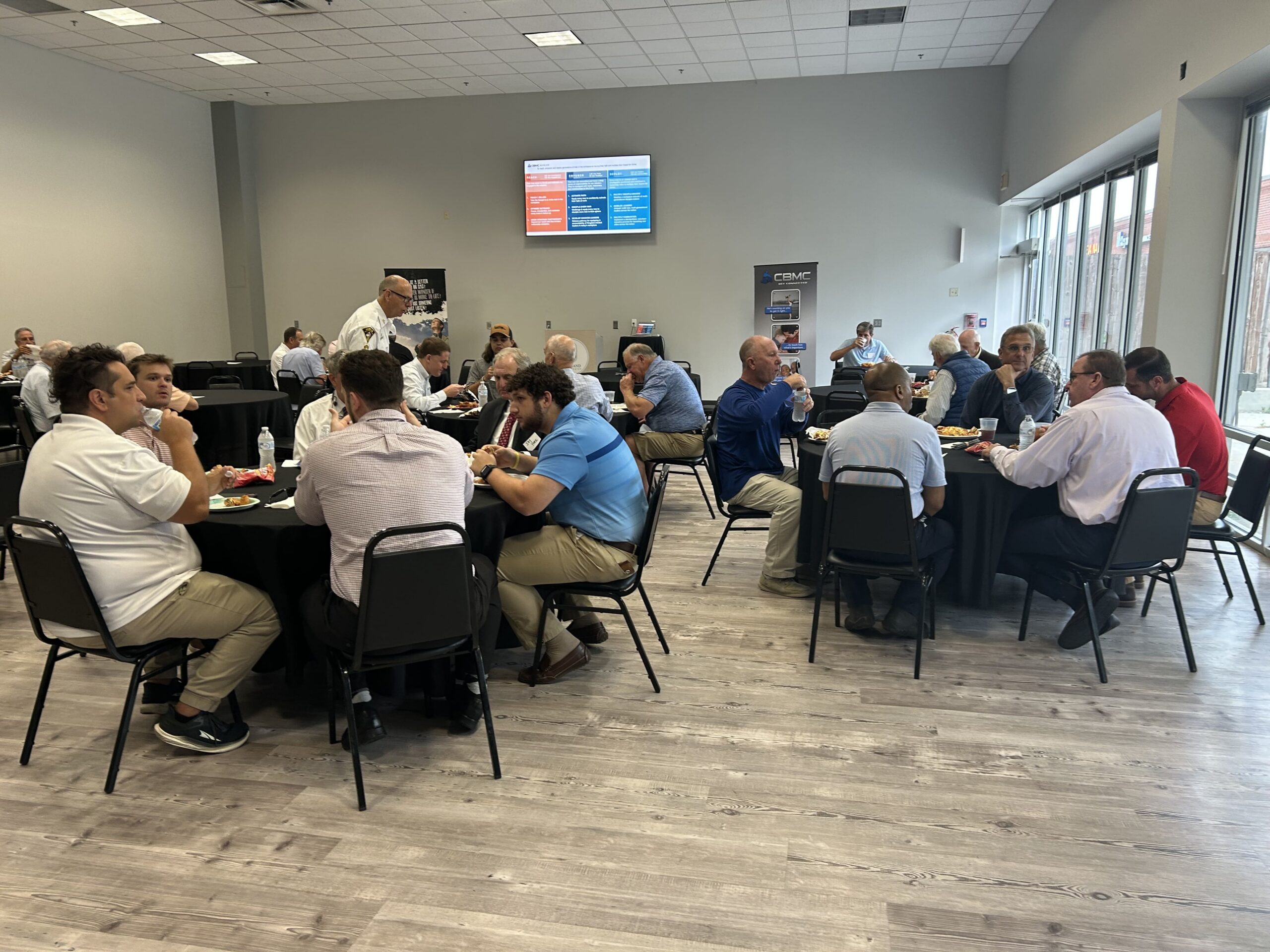 Attendees converse and eat at round tables in a modern conference room with large windows and a front-facing information screen.