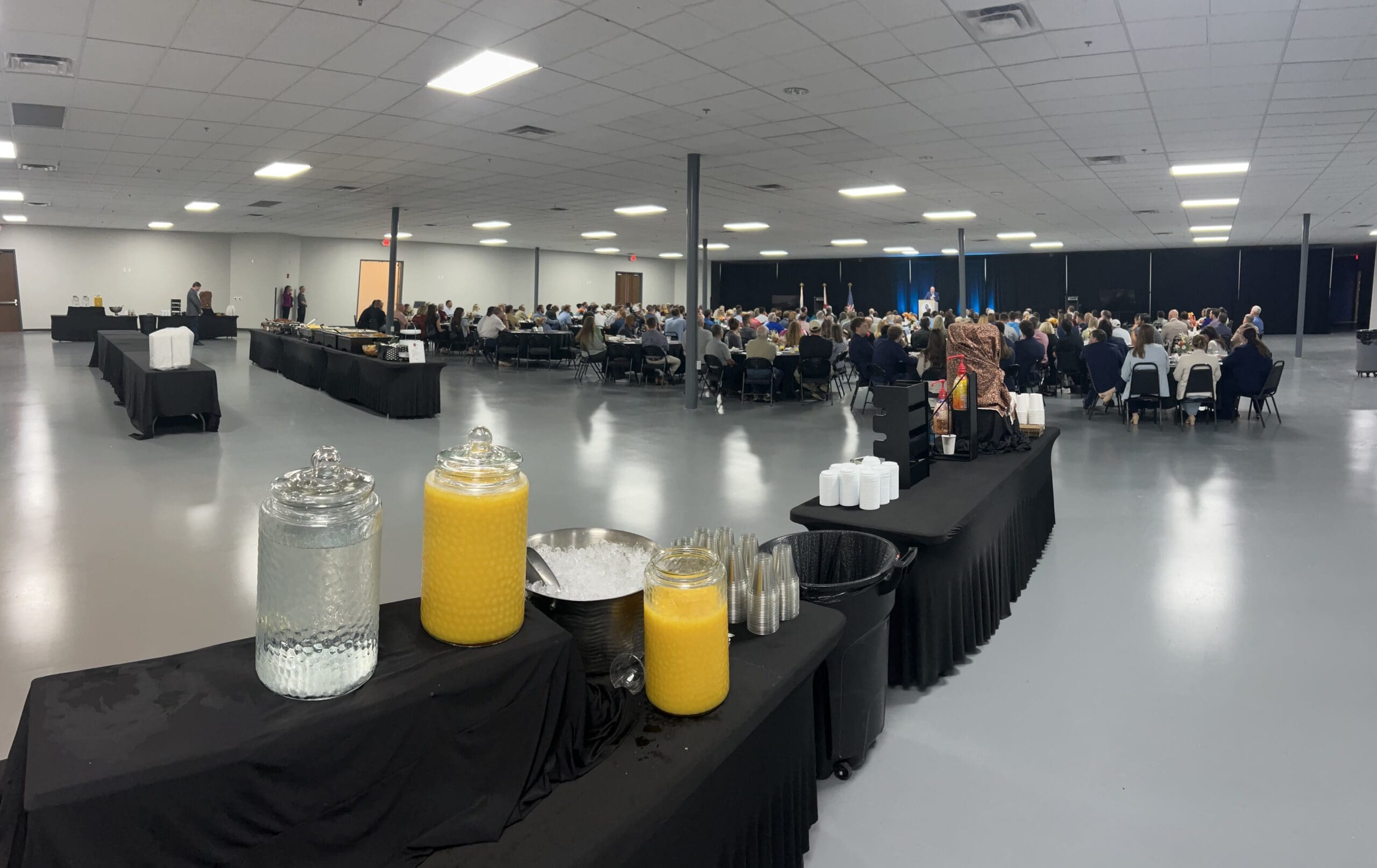 Event in spacious room with seated guests; foreground shows refreshment table with juice, water, cups on gray floor.