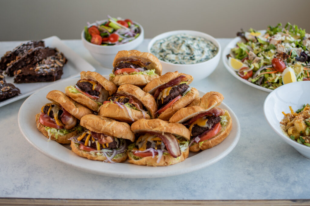 Croissant sandwiches with meat, cheese, and veggies are served on a platter with salad bowls, dip, and brownies nearby.