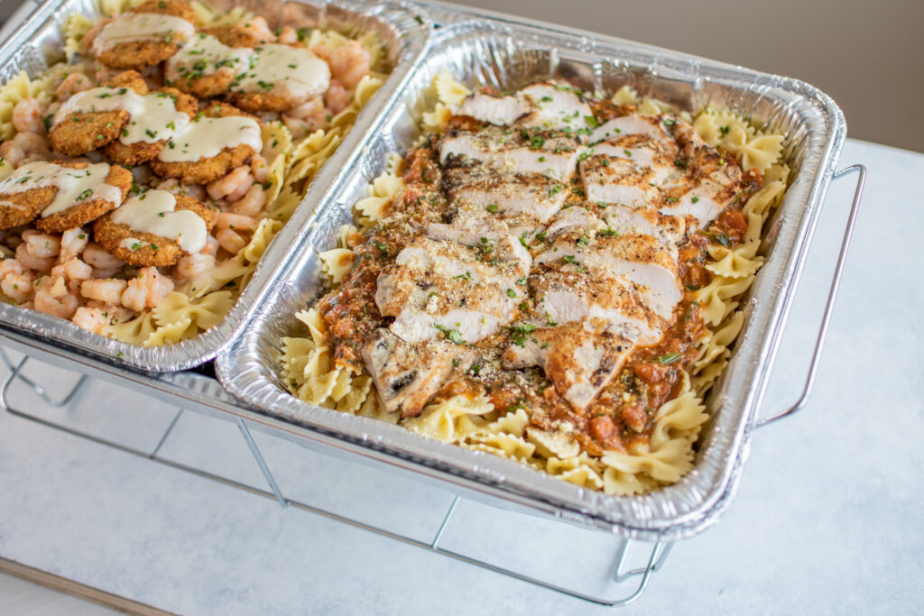 Two foil pasta trays: one with breaded eggplant and shrimp in creamy sauce, one with sliced chicken and tomato sauce, garnished with herbs.