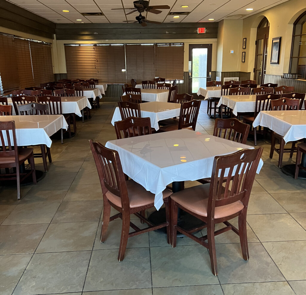Copeland's of New Orleans - Houma Banquet Room with wooden chairs, square tables with white tablecloths, tiled floor, and brown window blinds.