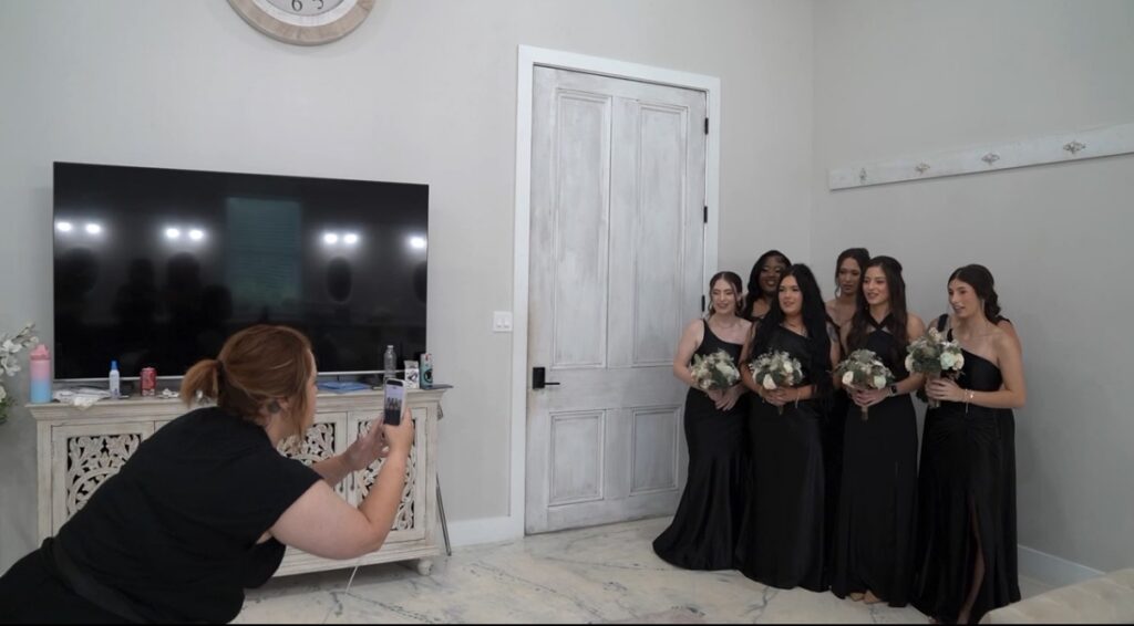 Anna snaps photo of six bridesmaids in black dresses with bouquets, posing indoors near a white door and TV, at Copeland’s Houma Catering.