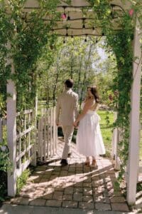 A couple holding hands walks through a garden trellis covered in vines.