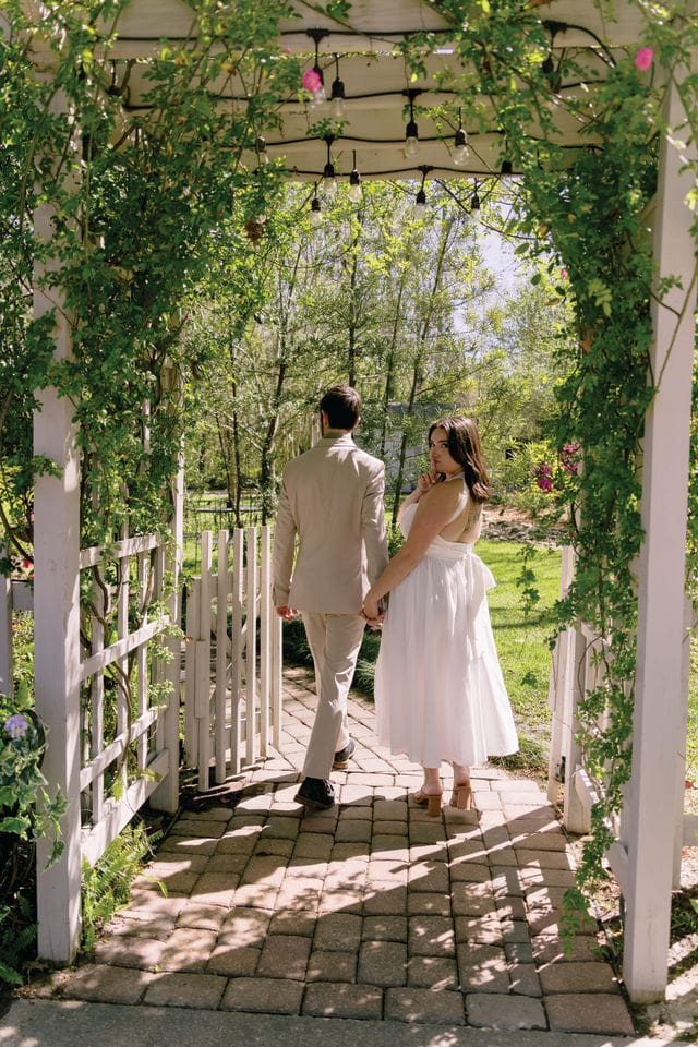A couple holding hands walks through a garden trellis covered in vines.