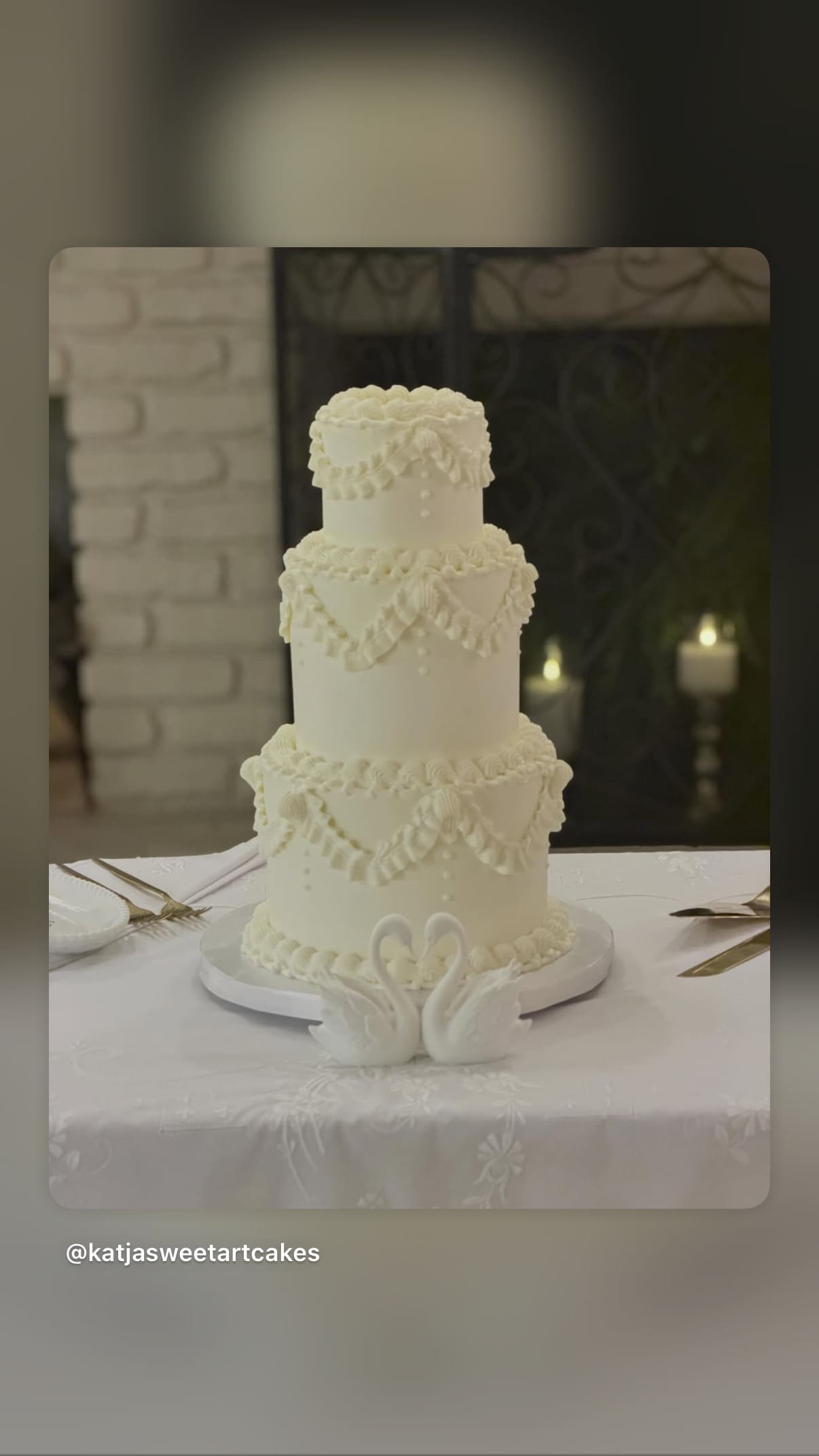 Three-tiered white wedding cake by SweetArt Cakes with piped icing and swan cake toppers on a table.