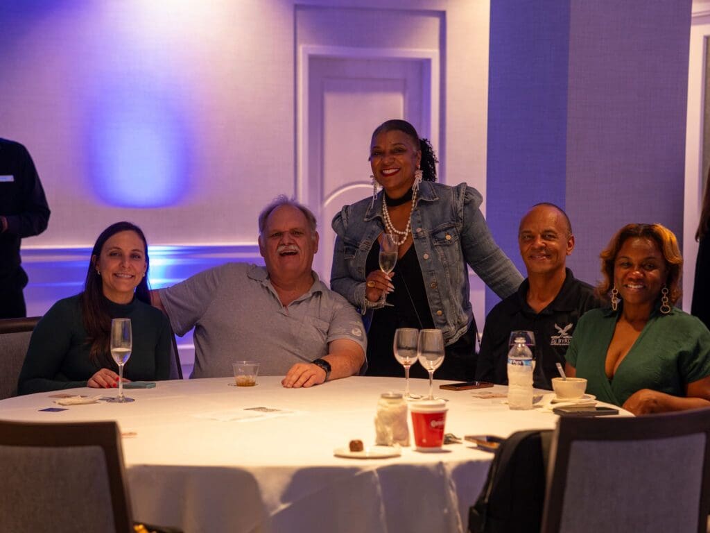 Five people smiling at a round event table with one person standing behind holding a glass.