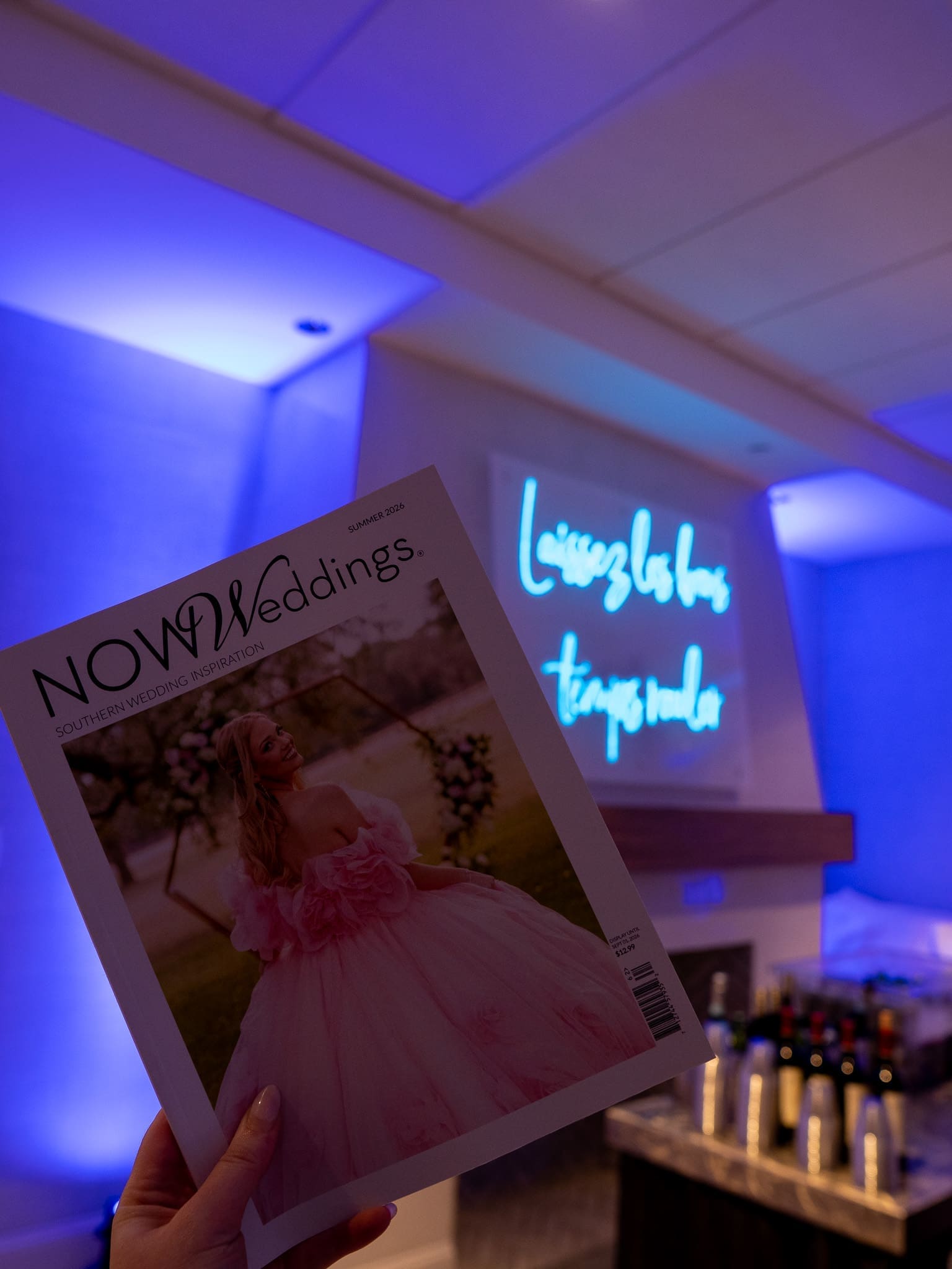 A hand holding NOW Weddings magazine in front of a blue neon sign.